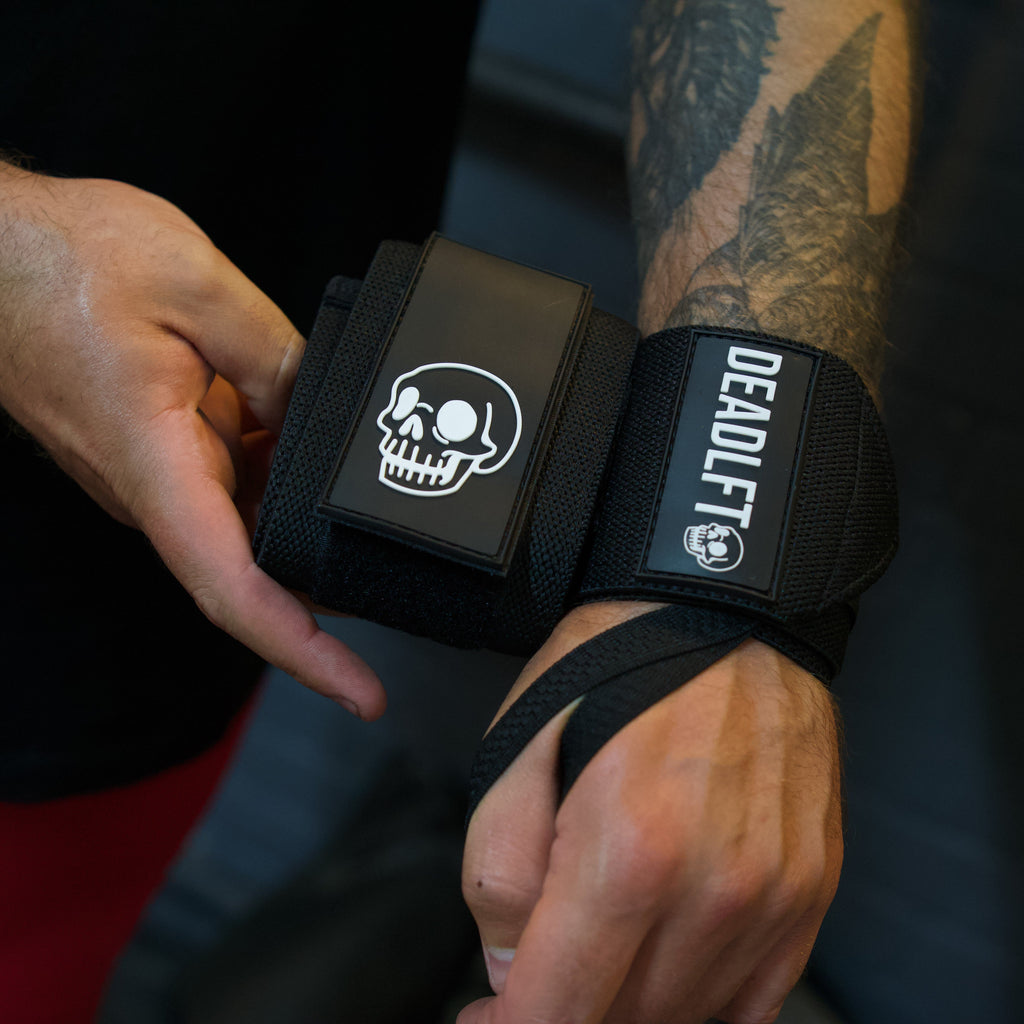 A close-up of a person's tattooed arm wearing a black Deadlift wrist wrap with a skull logo and white branding, while another hand adjusts the strap.