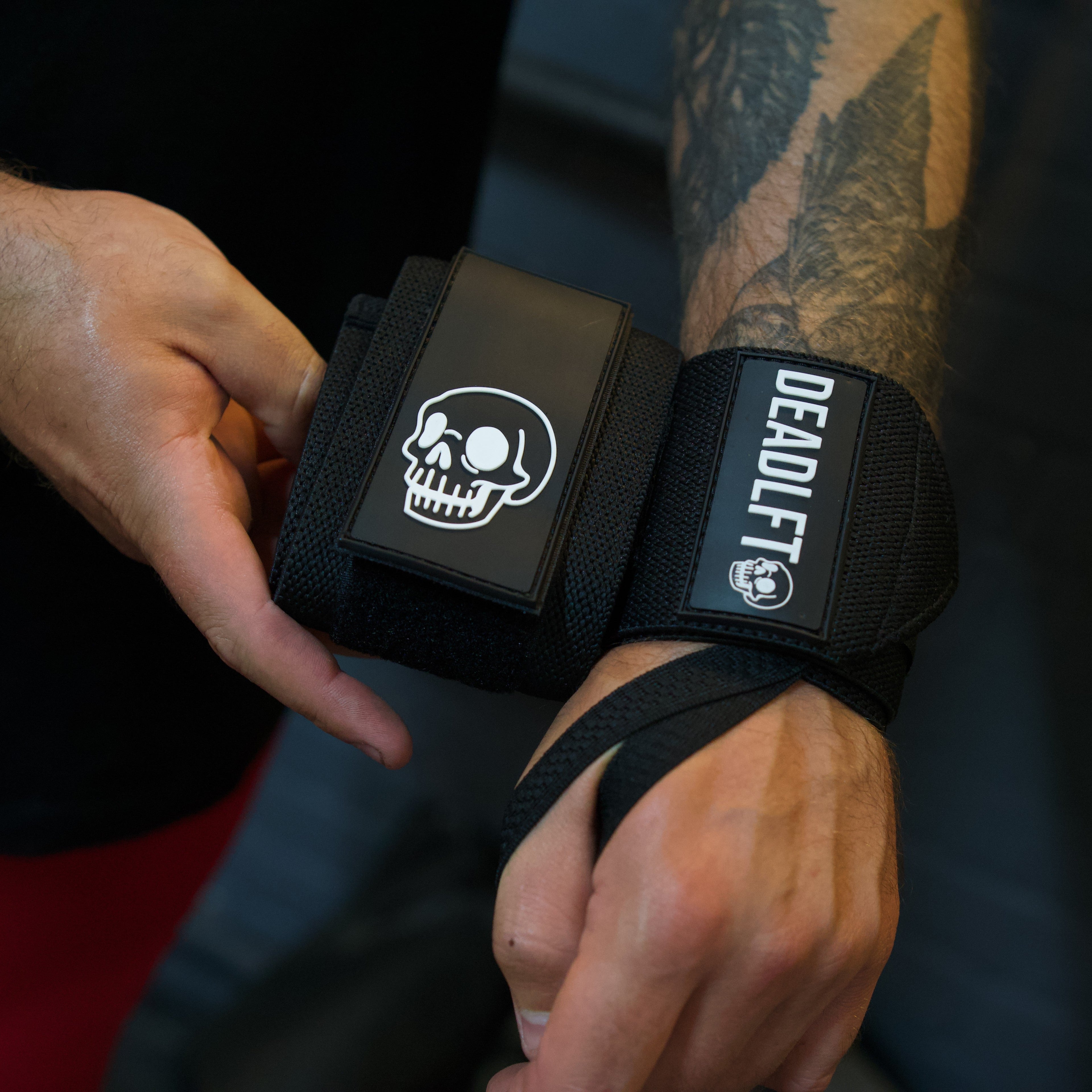 A close-up of a person's tattooed arm wearing a black Deadlift wrist wrap with a skull logo and white branding, while another hand adjusts the strap.