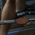A close-up of a person's tattooed arm and hand gripping a barbell in a gym, wearing a black Deadlift wrist wrap.