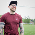 Man wearing a maroon t-shirt and black cap standing on a grassy field.